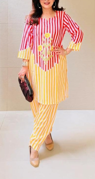A woman modeling a two-piece kurta set with red and white vertical stripes on the top and yellow and white vertical stripes on the pants, holding a small black clutch in a tiled indoor setting.