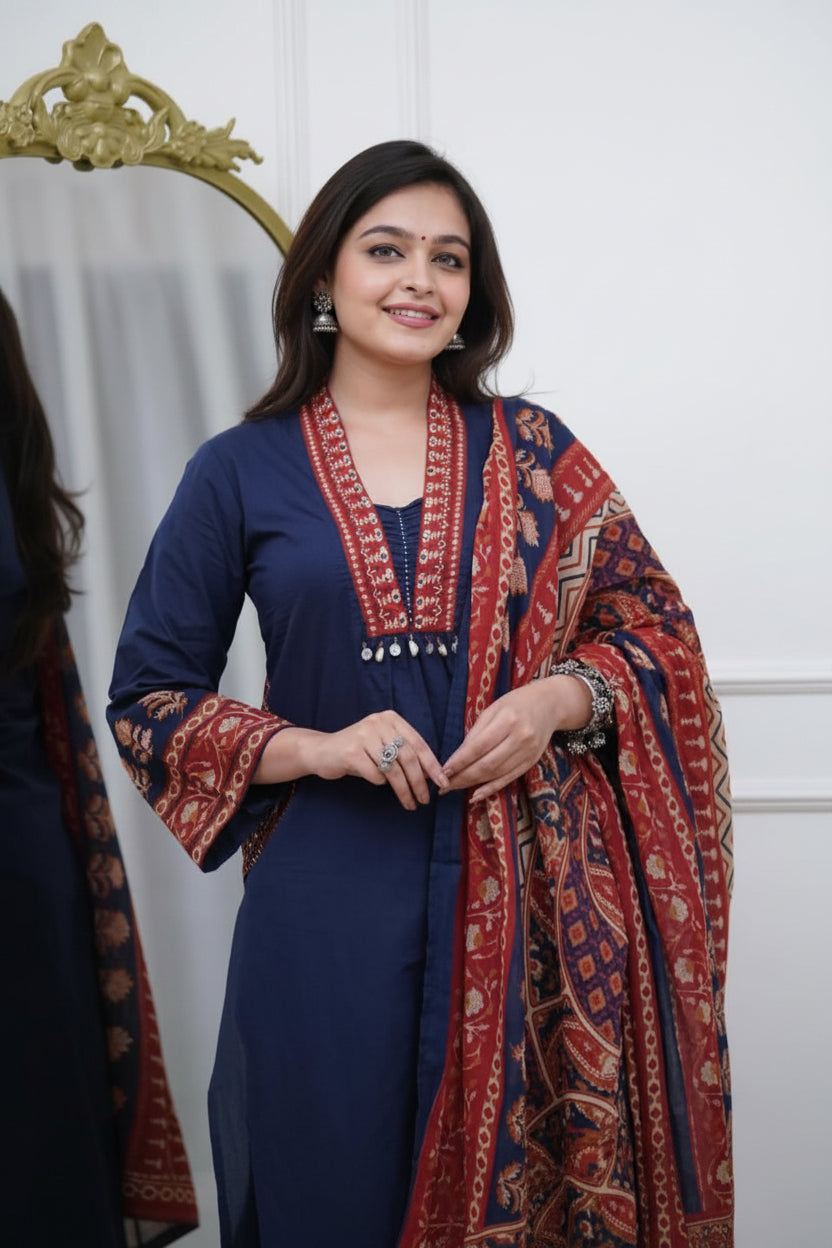 Woman wearing a blue outfit with a patterned red and orange scarf, standing in front of a white curtain.