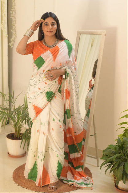 A woman wearing a white saree with orange and green geometric patterns and a matching orange blouse, standing indoors next to a mirror with potted plants in the background.