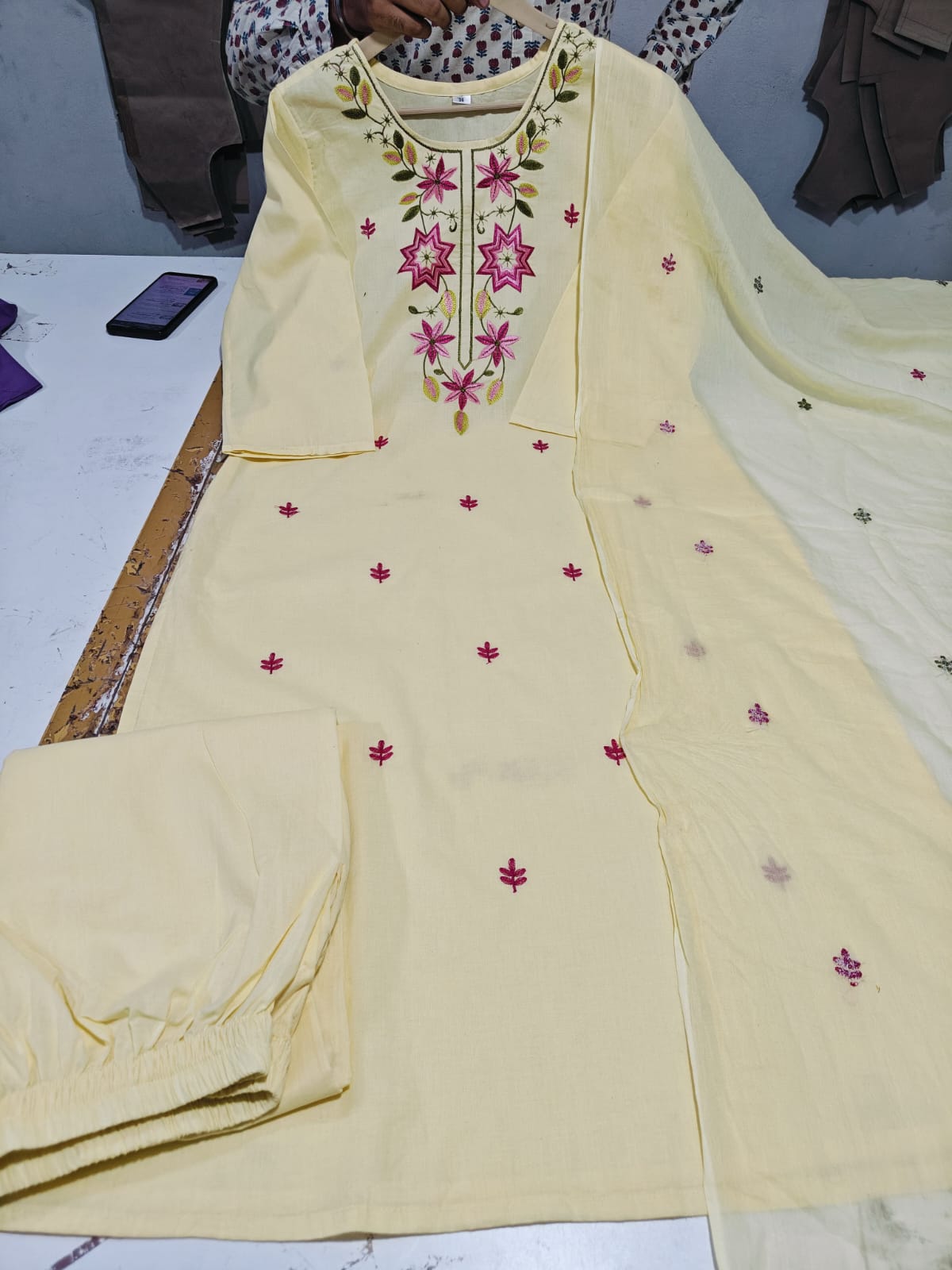 A light yellow ethnic salwar kameez suit set with matching pants and dupatta, featuring pink floral embroidery on the neckline and scattered pink motifs on the fabric.