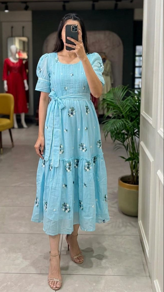 A woman takes a mirror selfie in a boutique wearing a light blue tiered midi dress with puff sleeves and a floral print. The dress features a tie waist and a square neckline.