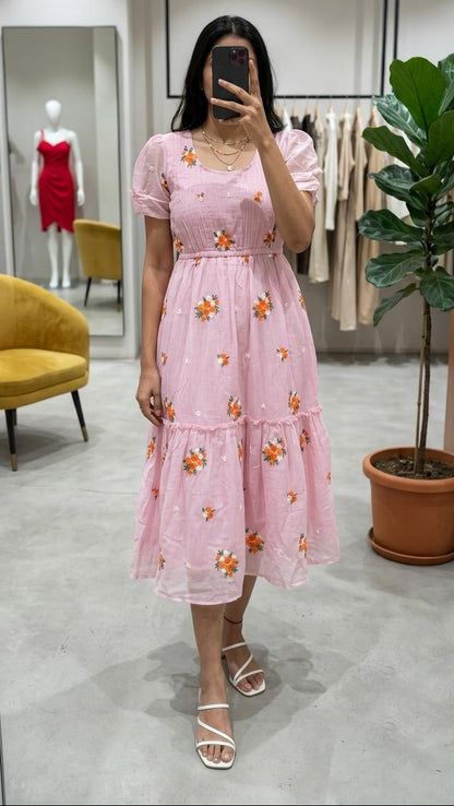A woman takes a mirror selfie in a boutique wearing a light pink tiered midi dress with orange floral embroidery and puff sleeves. The boutique features a gray floor, a yellow velvet chair, a potted fiddle-leaf fig plant, and a mannequin displaying a red dress in the background.