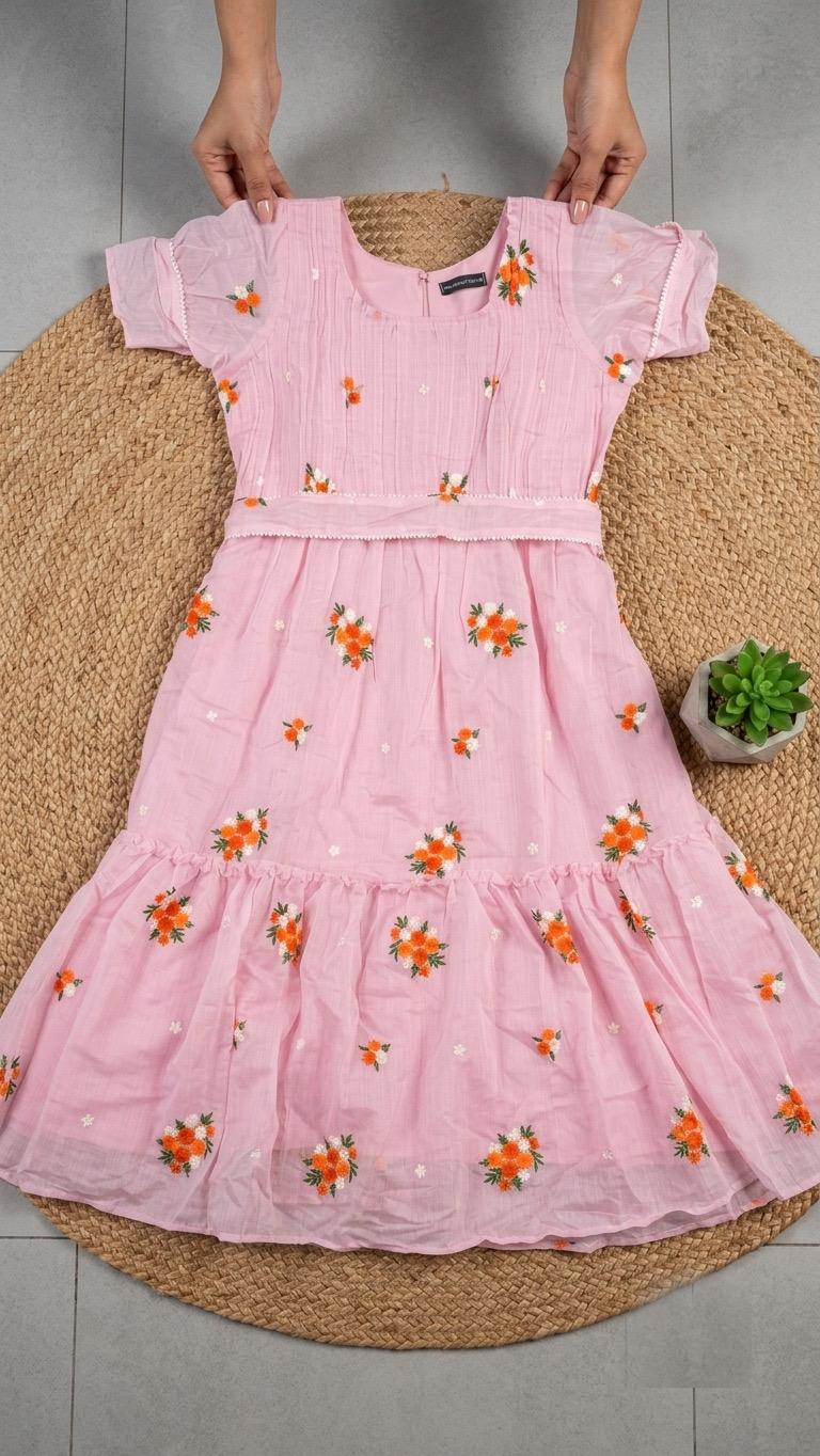 A pink short-sleeve cotton dress with orange and yellow floral embroidery, held up by hands over a round woven jute mat.