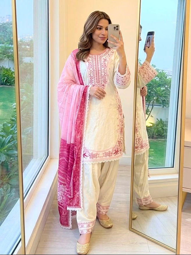 A woman takes a mirror selfie in a modern room wearing a traditional Indian salwar kameez outfit. The outfit consists of a cream-colored embroidered kurta with a pink border, cream-colored pants, and a pink dupatta draped over her shoulder.