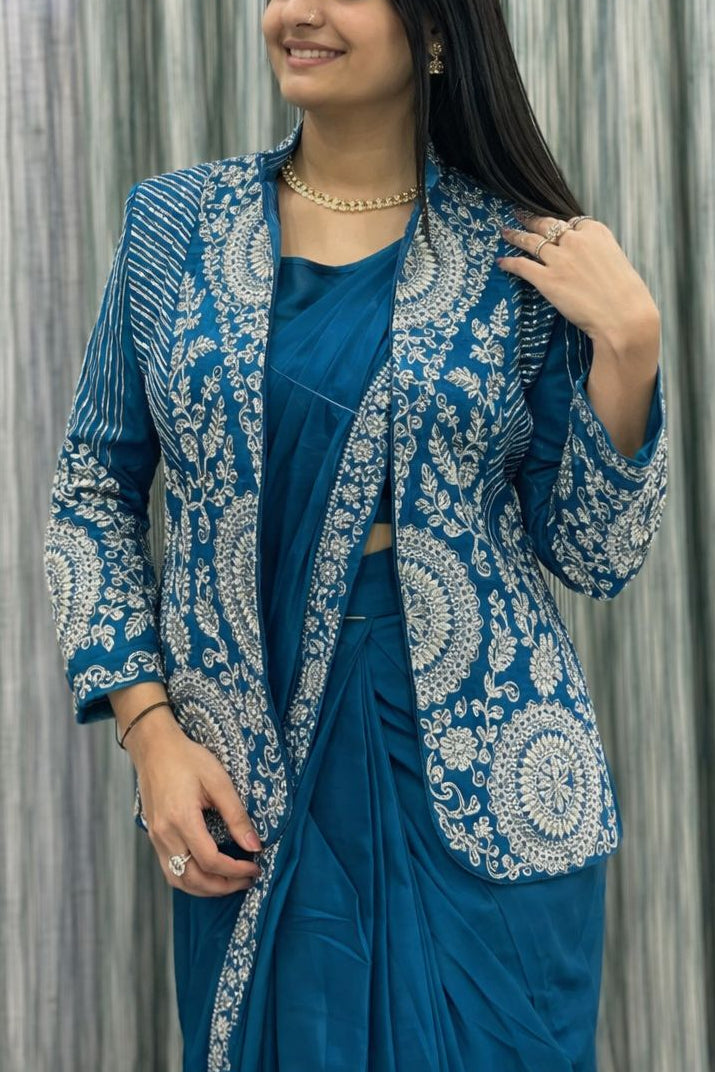 Woman wearing a blue and white embroidered jacket over a blue saree against a textured wall.