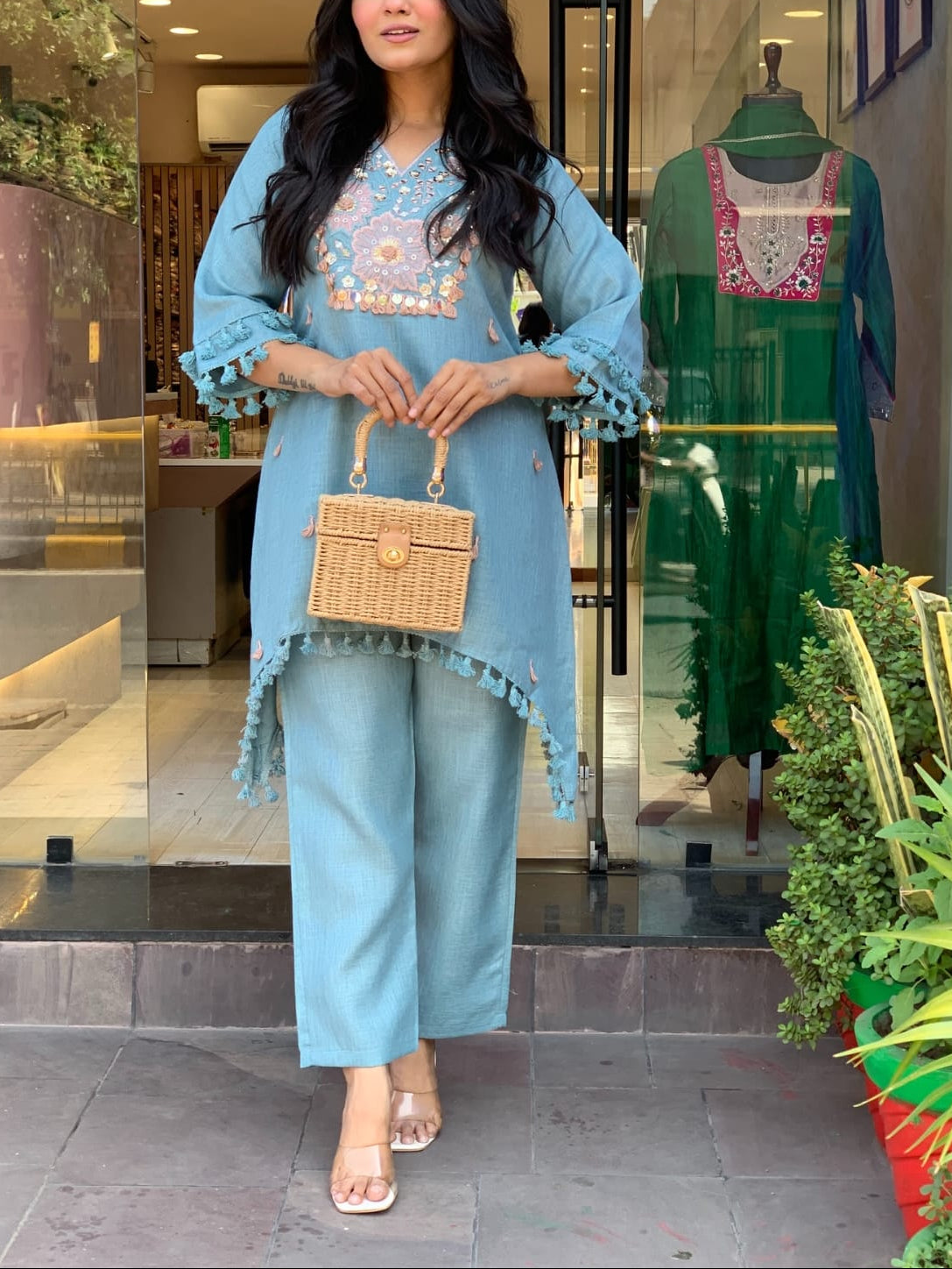 A woman stands in front of a boutique window wearing a light blue embroidered tunic and matching wide-leg pants, holding a small woven handbag. A green outfit is visible on a mannequin inside the shop, with plants along the storefront.