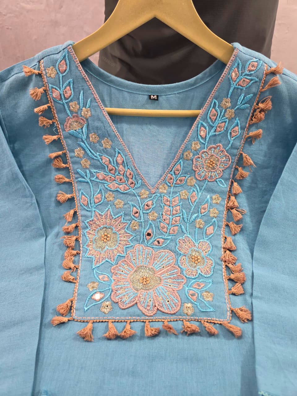 A light blue cotton kurta with intricate peach and gold floral embroidery and tassels on the neckline, hanging on a wooden hanger.