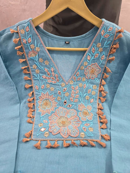 A light blue cotton kurta with intricate peach and gold floral embroidery and tassels on the neckline, hanging on a wooden hanger.