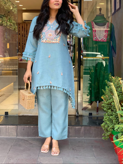 A woman models a light blue three-piece kurta set with floral embroidery and tassel trim, standing in front of a clothing store window.