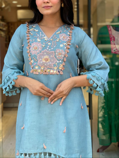 A woman wearing a light blue embroidered kurta with floral patterns and tassel trim, standing in front of a glass storefront.