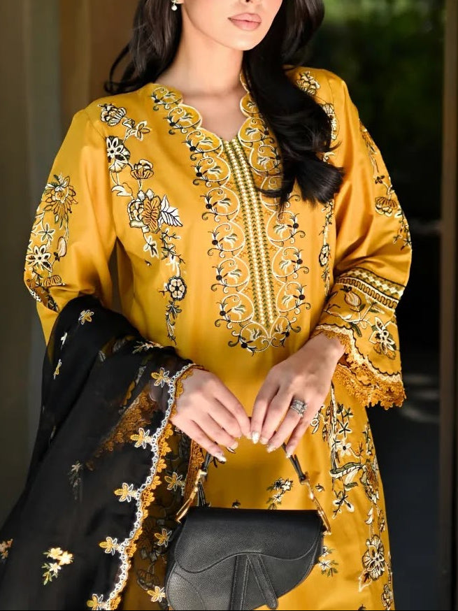 A woman models a traditional three-piece salwar kameez suit in mustard yellow with intricate black and gold floral embroidery, paired with a black dupatta featuring gold embroidery, set against an arched stone doorway.