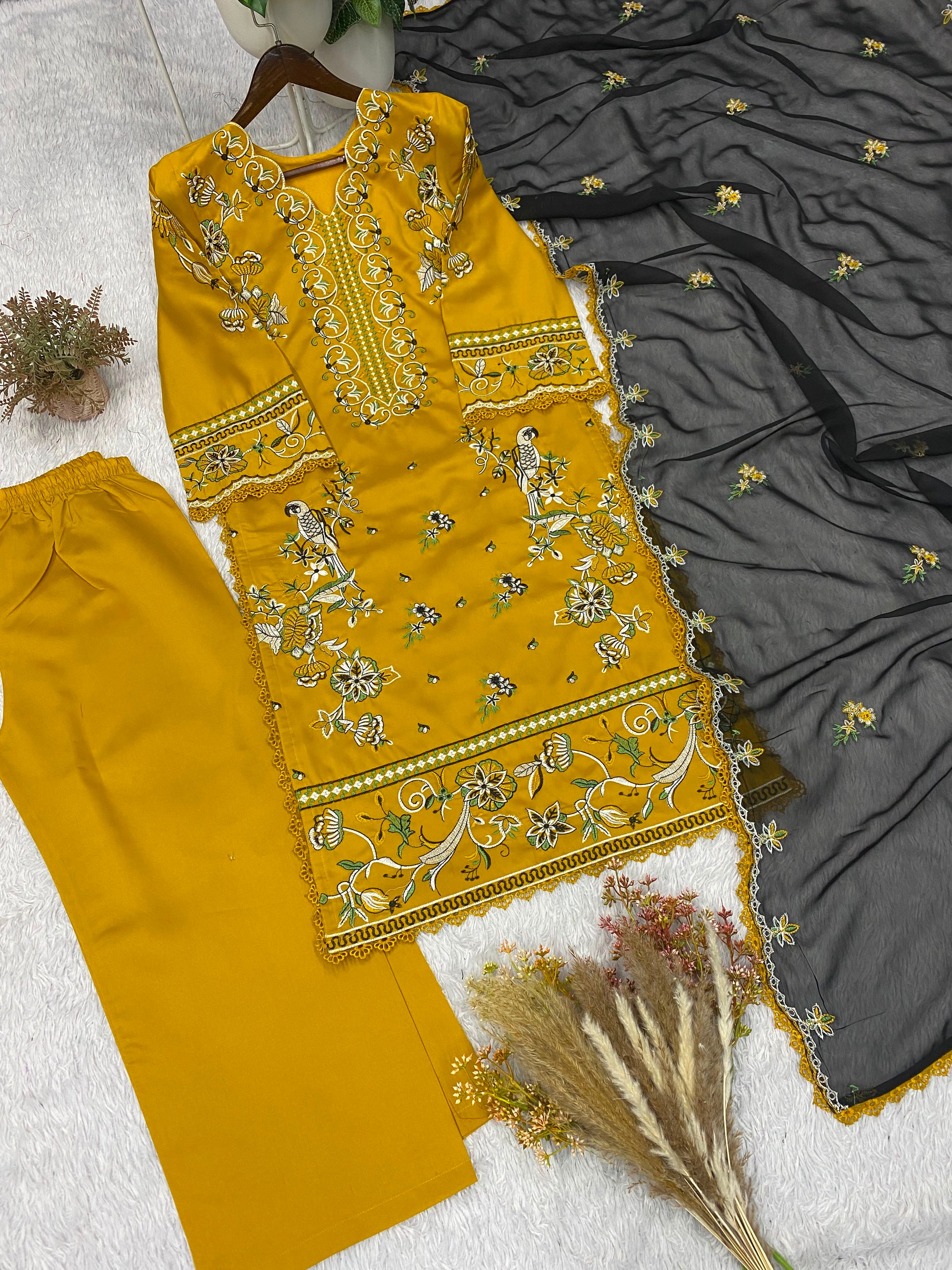 A three-piece women's ethnic suit set laid out on a white textured surface, featuring a mustard yellow embroidered kurta, matching yellow trousers, and a dark gray embroidered dupatta.