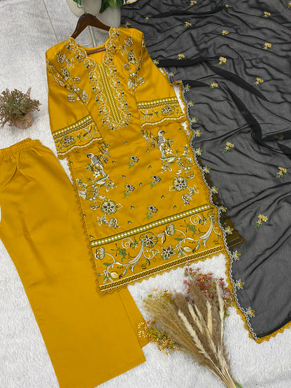 A three-piece women's ethnic suit set laid out on a white textured surface, featuring a mustard yellow embroidered kurta, matching yellow trousers, and a dark gray embroidered dupatta.