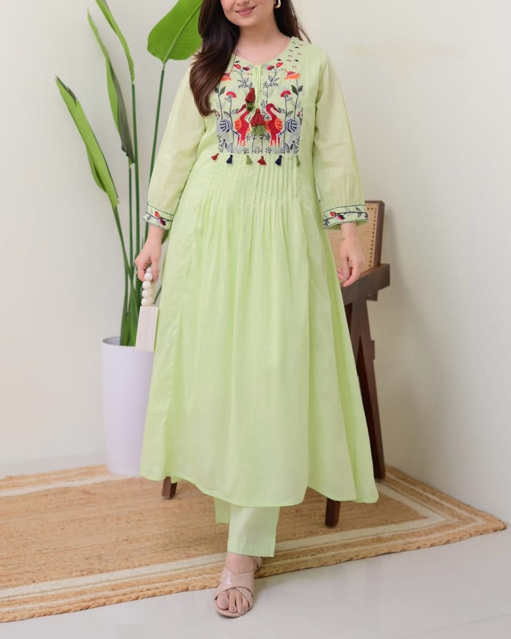 Woman wearing a light green embroidered dress standing indoors with a plant and chair in the background.