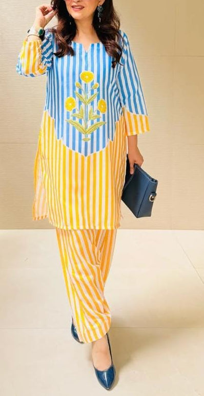 A woman modeling a two-piece kurta set with blue and yellow vertical stripes, featuring a yellow floral embroidered design on the chest and matching striped pants, set against a beige tiled wall.