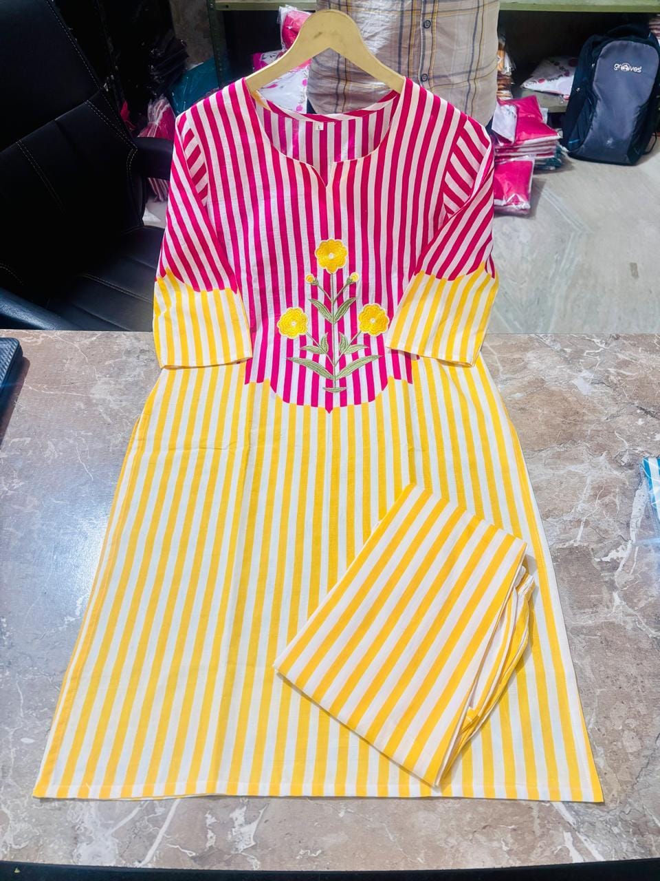 A women's kurta set with matching pants, featuring a vibrant pink and yellow vertical stripe pattern, a yellow yoke with yellow floral embroidery, and a folded pair of matching striped pants on a wooden hanger, displayed on a marble countertop.