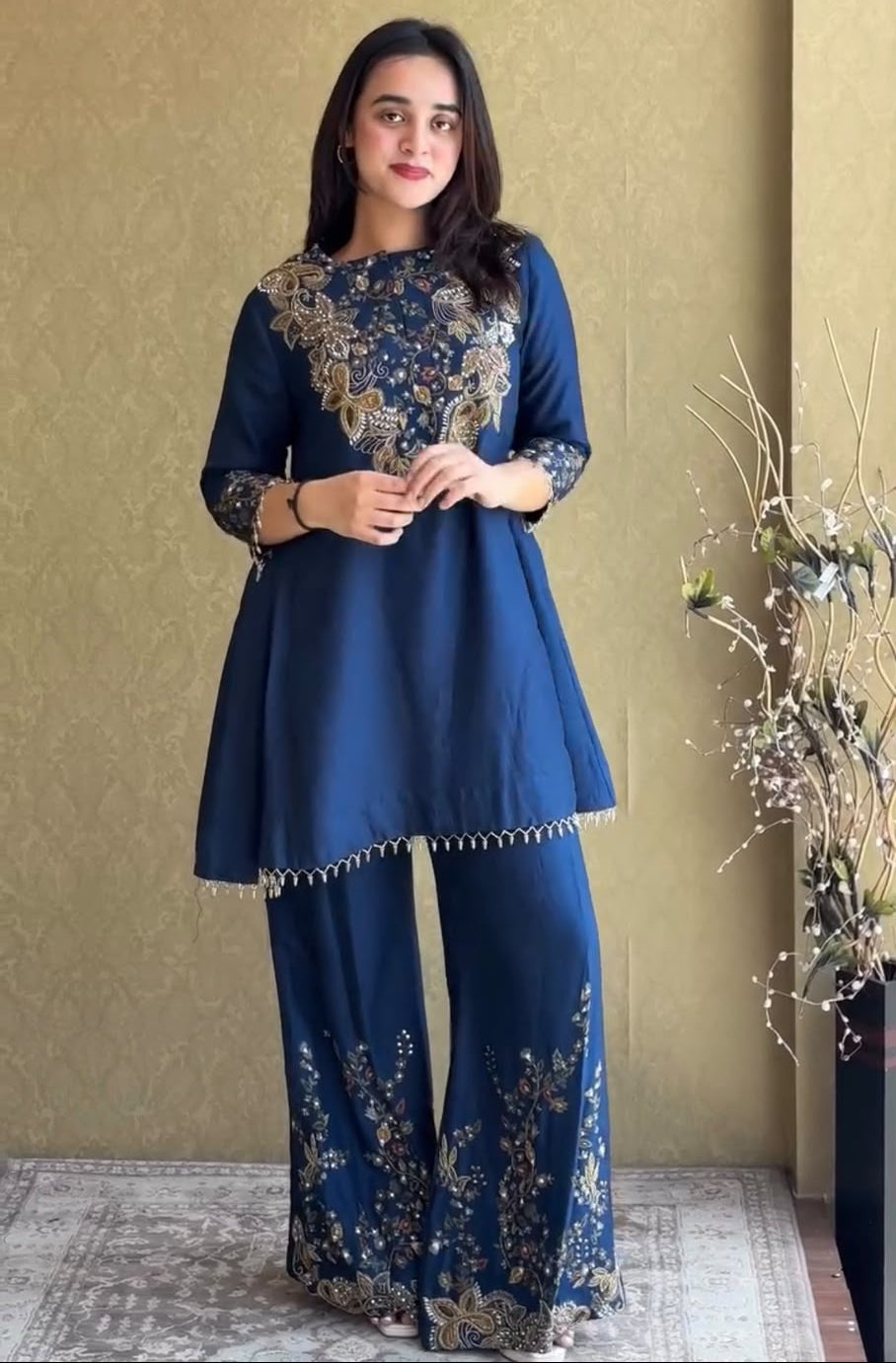 A woman modeling a navy blue traditional outfit with intricate gold embroidery on the kurta and wide-leg pants, standing against a textured beige wall.