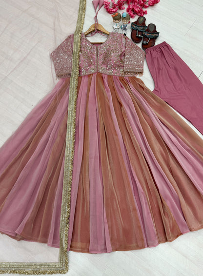 A pink and rose anarkali-style dress set laid flat on a white fabric background, featuring a heavily embellished pink bodice, a layered tulle skirt in pink and dusty rose tones, matching pants, and a gold-trimmed dupatta. Small decorative shoes and flowers are placed around the outfit.