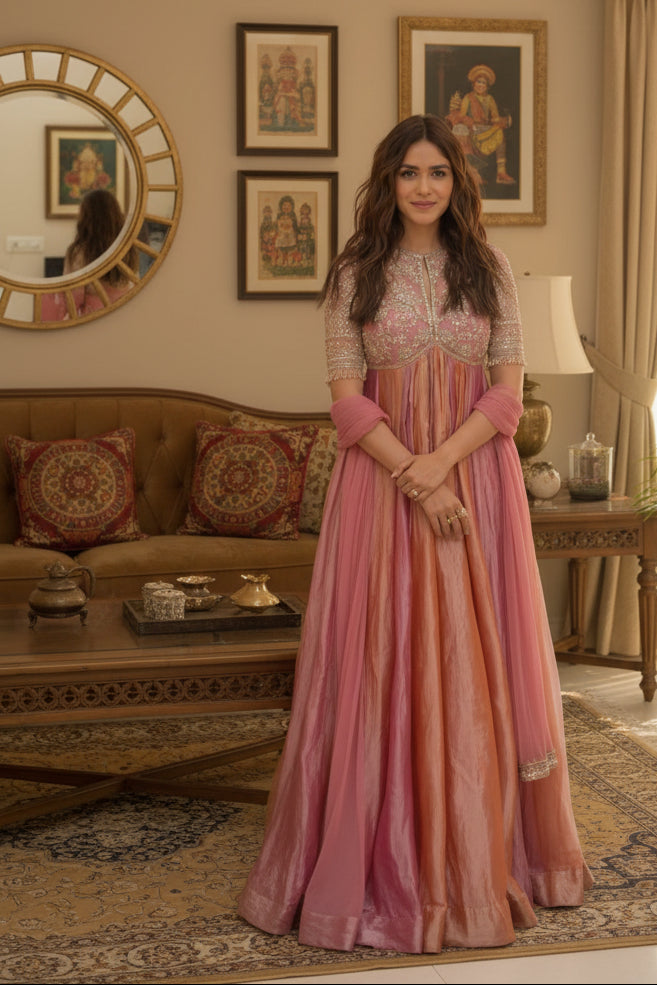 A woman wearing a long pink and peach embroidered anarkali-style gown stands outdoors with her hands clasped in front of a motorcycle and construction backdrop.
