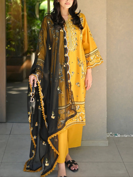 A woman models a traditional three-piece salwar kameez suit in mustard yellow and black with gold embroidery, standing in front of a beige stucco archway.